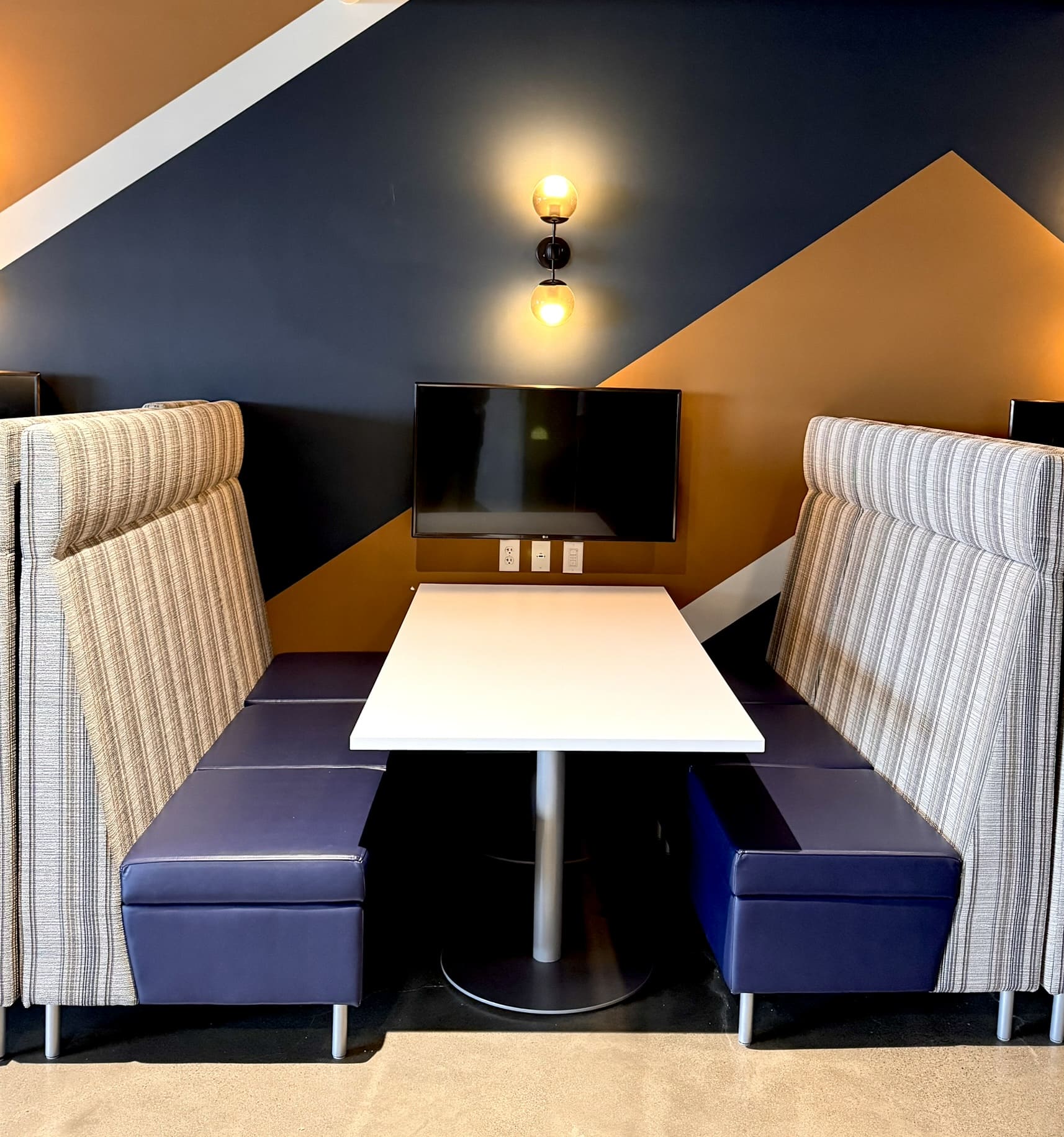 Close-up of a banquette seating area with two high-back upholstered benches, blue leather seats, a white rectangular table, a wall-mounted TV, and a double globe sconce light against a navy and gold geometric accent wall.
