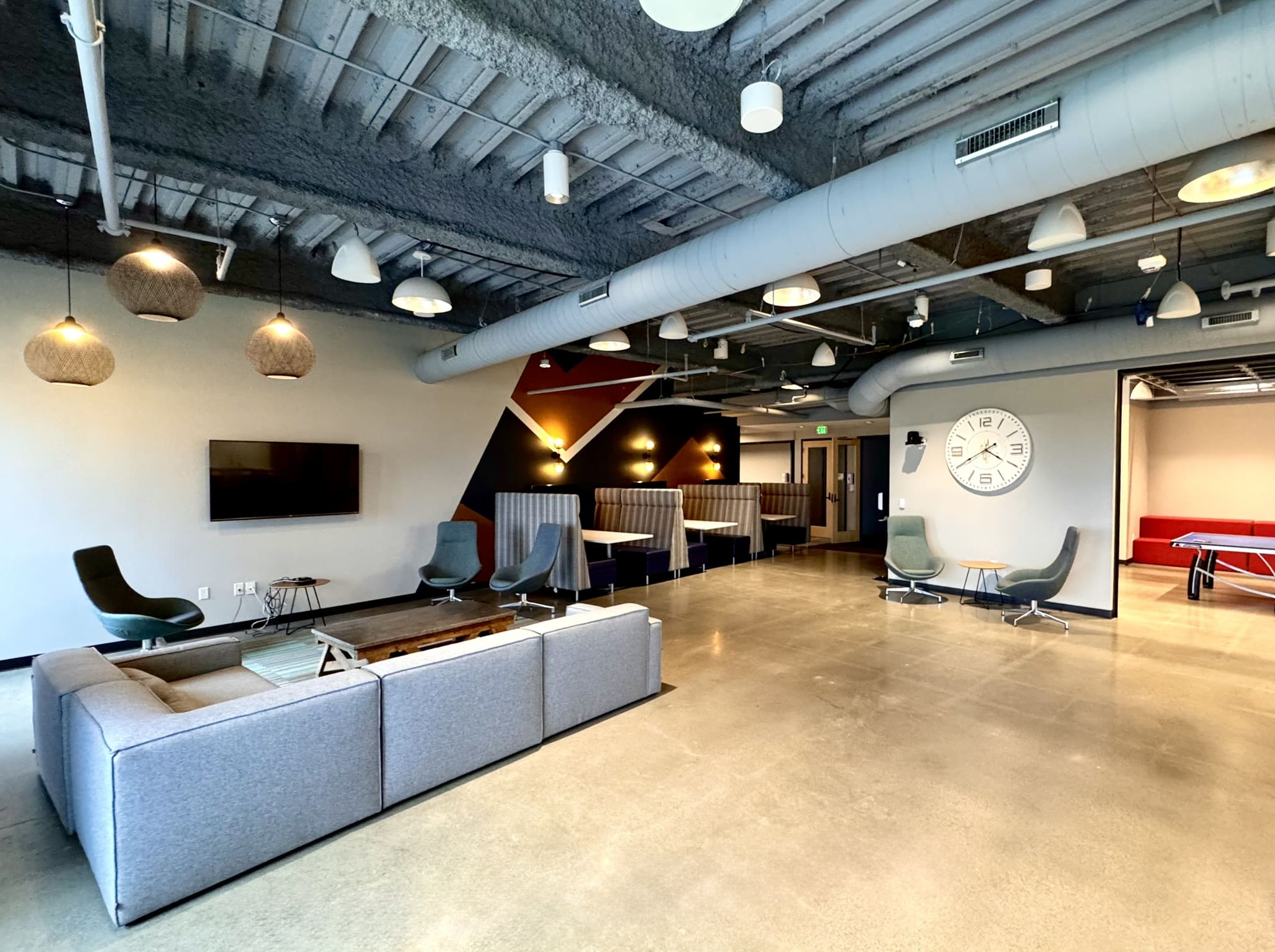 Spacious open common area with a gray sectional sofa, lounge chairs, banquette booths with high-back seating, a wall-mounted TV, woven pendant lights, a large wall clock, and a ping pong table visible in the background gaming area.