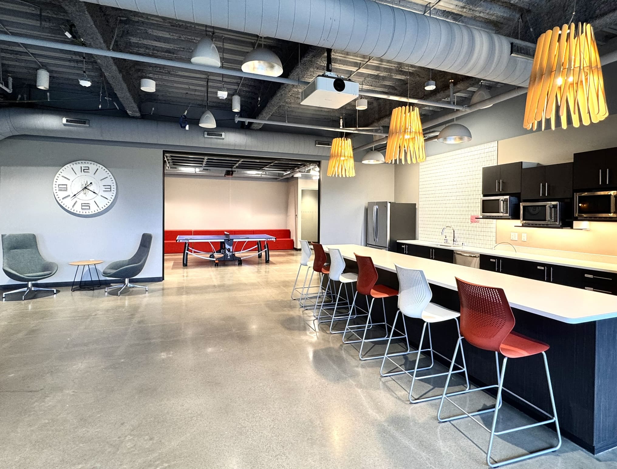Wide view of a modern open common area featuring a kitchen counter with red and white bar stools, wooden pendant lights, a ping pong table with red bench seating in the background, two lounge chairs with a side table, and a large wall clock on exposed concrete walls.