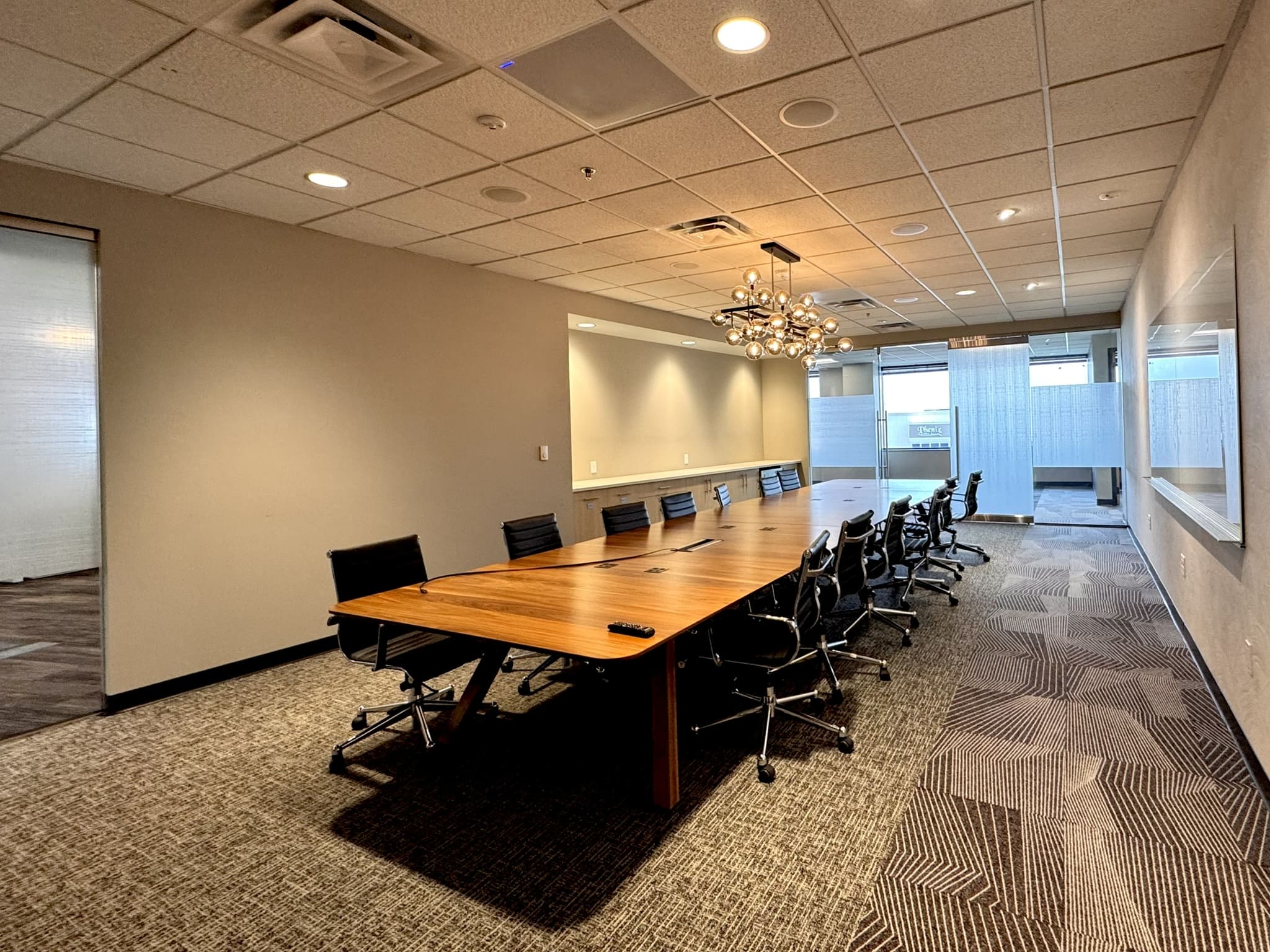 Large conference room with a long wooden table surrounded by black leather chairs, a modern multi-globe chandelier overhead, a projection screen on the back wall, and glass partition walls allowing natural light from an adjacent space.