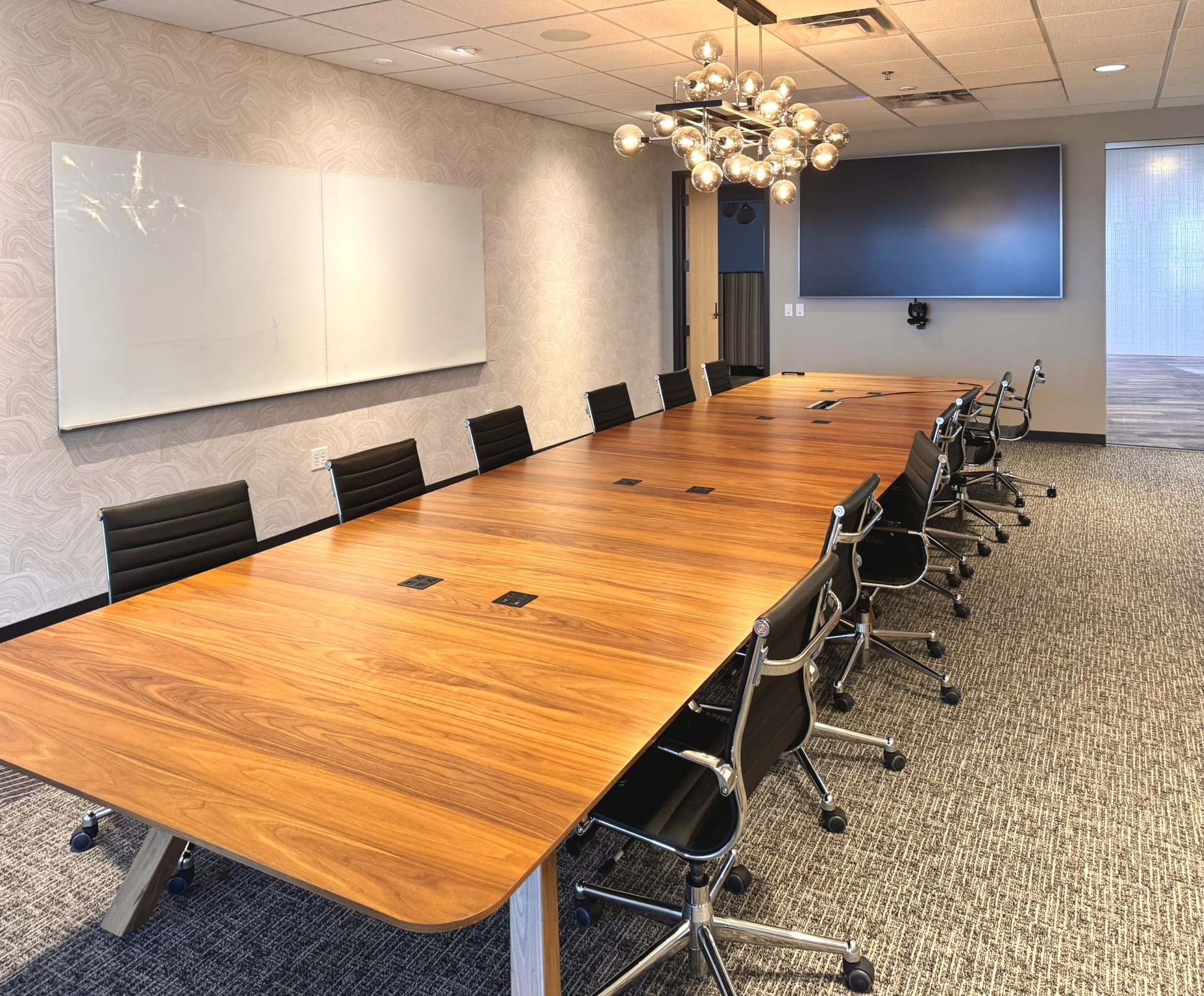 Elegant conference room with a long wooden table surrounded by black leather chairs, a large whiteboard on a textured accent wall, a modern multi-globe chandelier, and a mounted display screen at the far end of the room.