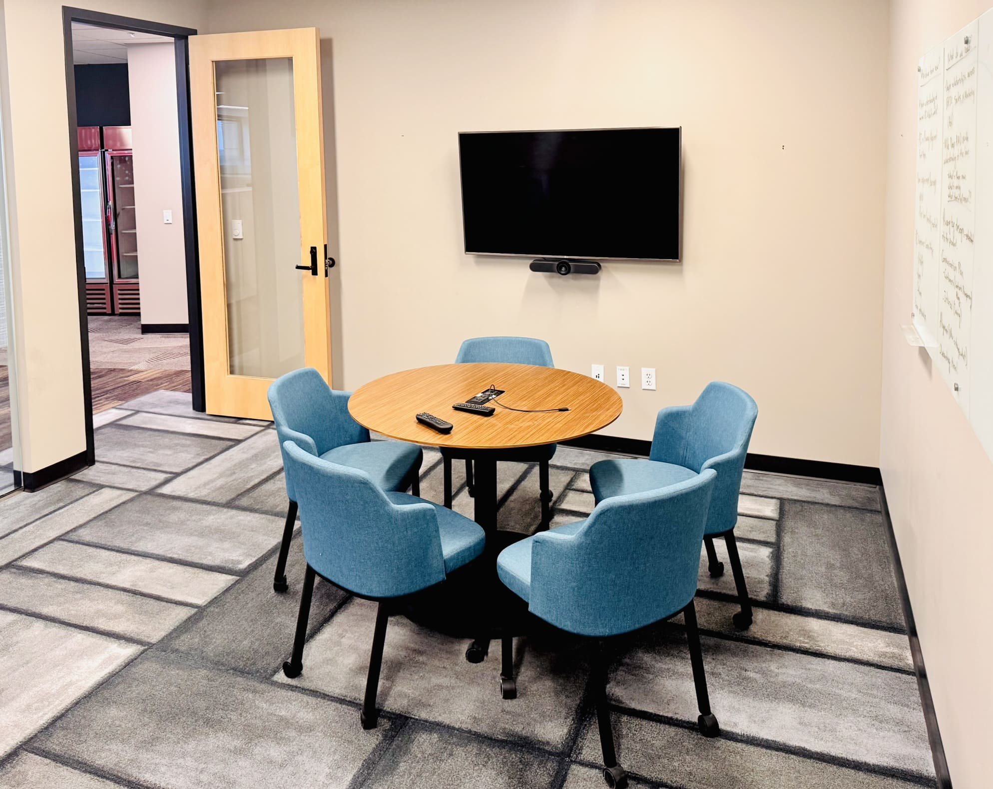 Small conference room with a round wooden table surrounded by four blue upholstered chairs, a wall-mounted TV with a video conferencing camera, a whiteboard on the right wall, and a glass-paneled wood door leading to the common area.