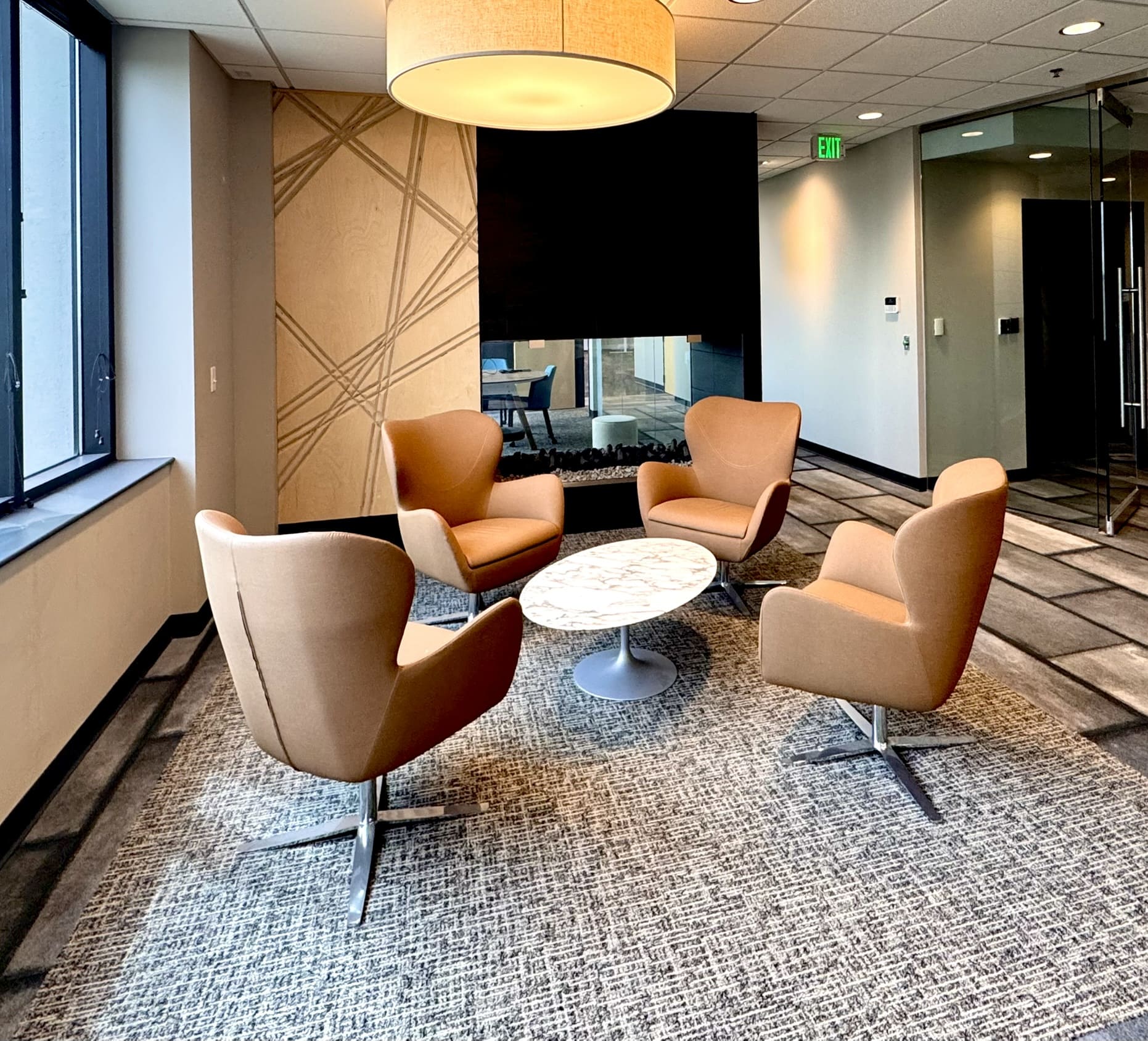 Lobby seating area with four tan leather swivel chairs arranged around a round marble-top table, a large drum pendant light overhead, a decorative geometric wood panel wall, and a fireplace in the background.