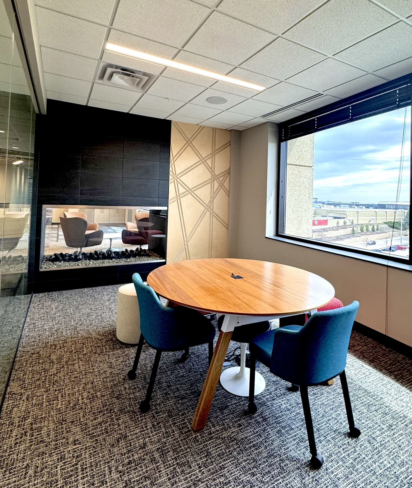 Small conference room with a round wooden table, three teal chairs and a round ottoman, a decorative geometric wood panel wall, a double-sided fireplace overlooking the lobby seating area, and a large window with an outdoor view.
